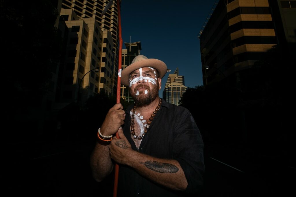 Image of Aboriginal artist, Tee Jay Worrigal. Tee Jay is holding a stick and wearing paint on his face as he stands on St Georges terrace